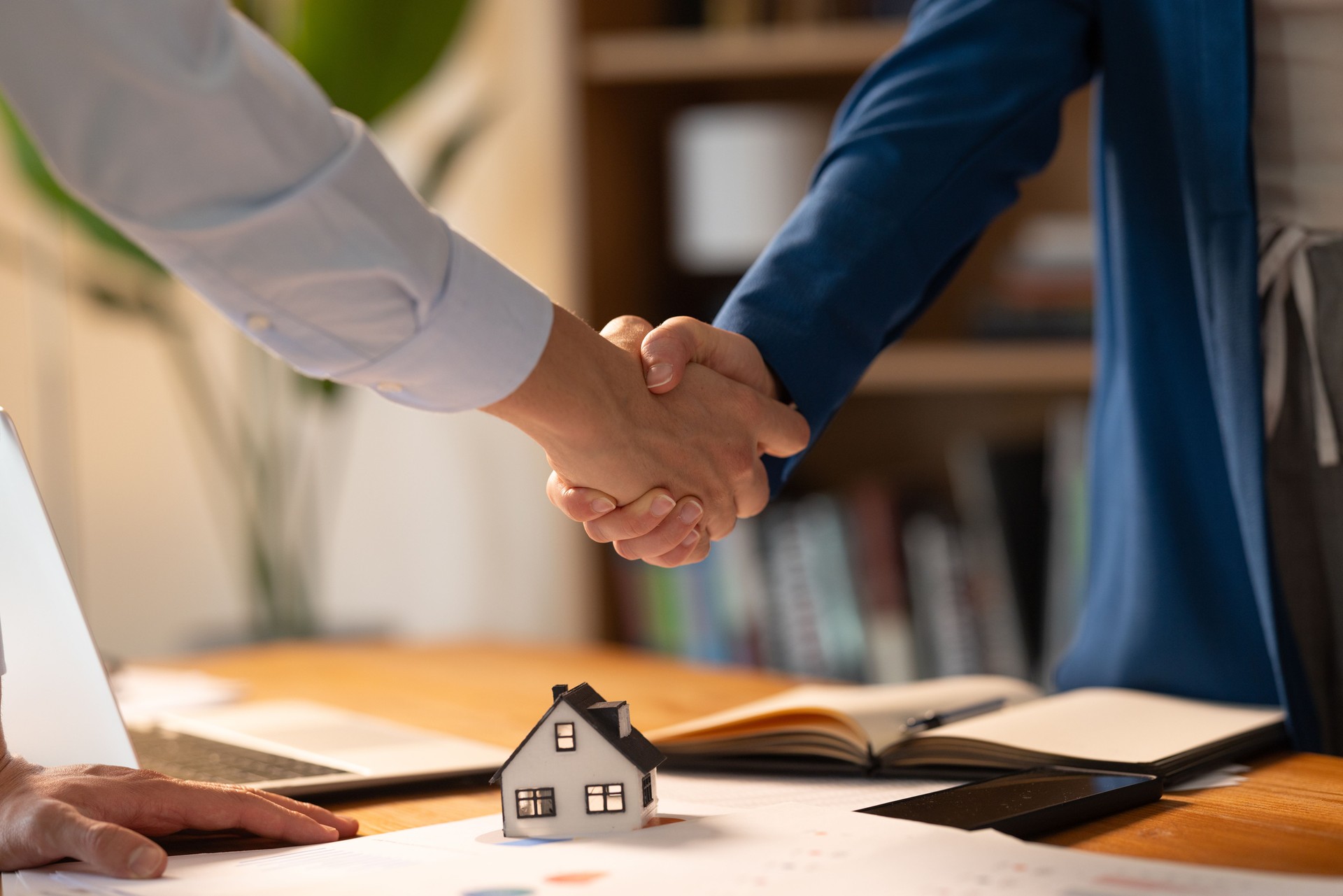Two people shaking hands over a miniature house model, closing a real estate deal